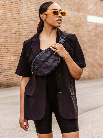 A stylish woman in a black blazer and biker shorts poses confidently outdoors with a black crossbody bag worn across her chest, accessorized with gold jewelry and amber sunglasses.