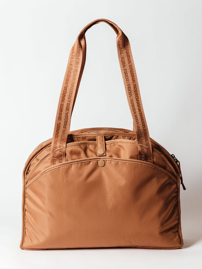  Front view of a structured caramel-colored duffel bag with a curved top, dual snap-closure front pockets, and wide shoulder straps featuring embroidered names in tonal thread.