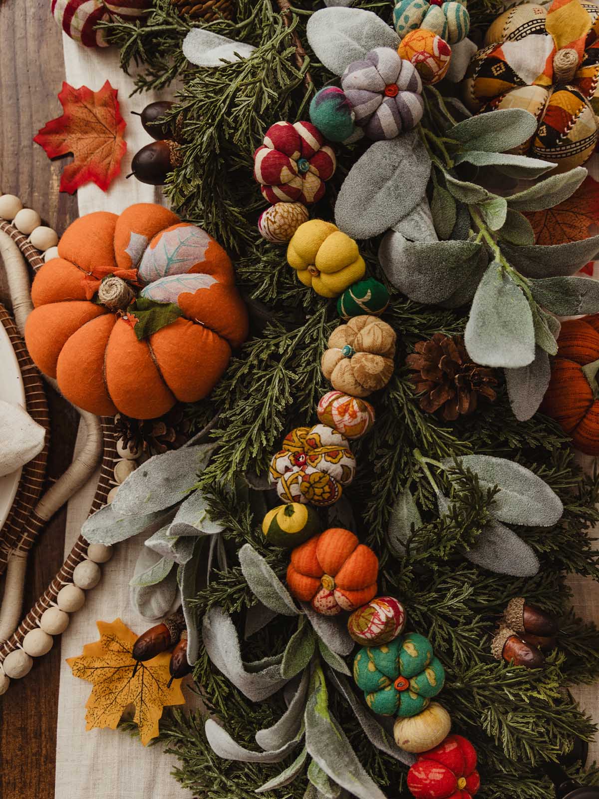 Top-down view of a lush fall centerpiece with greenery, faux lamb’s ear, pinecones, acorns, and a garland of small patterned fabric pumpkins in vibrant colors, anchored by larger fabric pumpkins at each end.