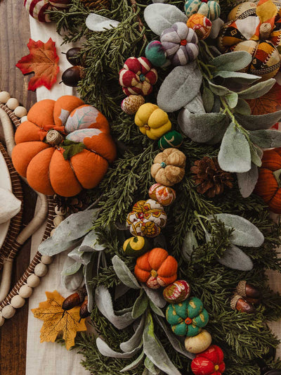 Top-down view of a lush fall centerpiece with greenery, faux lamb’s ear, pinecones, acorns, and a garland of small patterned fabric pumpkins in vibrant colors, anchored by larger fabric pumpkins at each end.