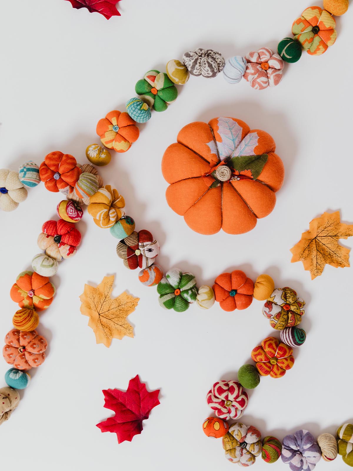 Colorful fabric pumpkin garland arranged on a white background with a large orange fabric pumpkin, scattered autumn leaves, and small patterned pumpkins in shades of green, orange, yellow, and red.