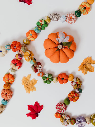 Colorful fabric pumpkin garland arranged on a white background with a large orange fabric pumpkin, scattered autumn leaves, and small patterned pumpkins in shades of green, orange, yellow, and red.
