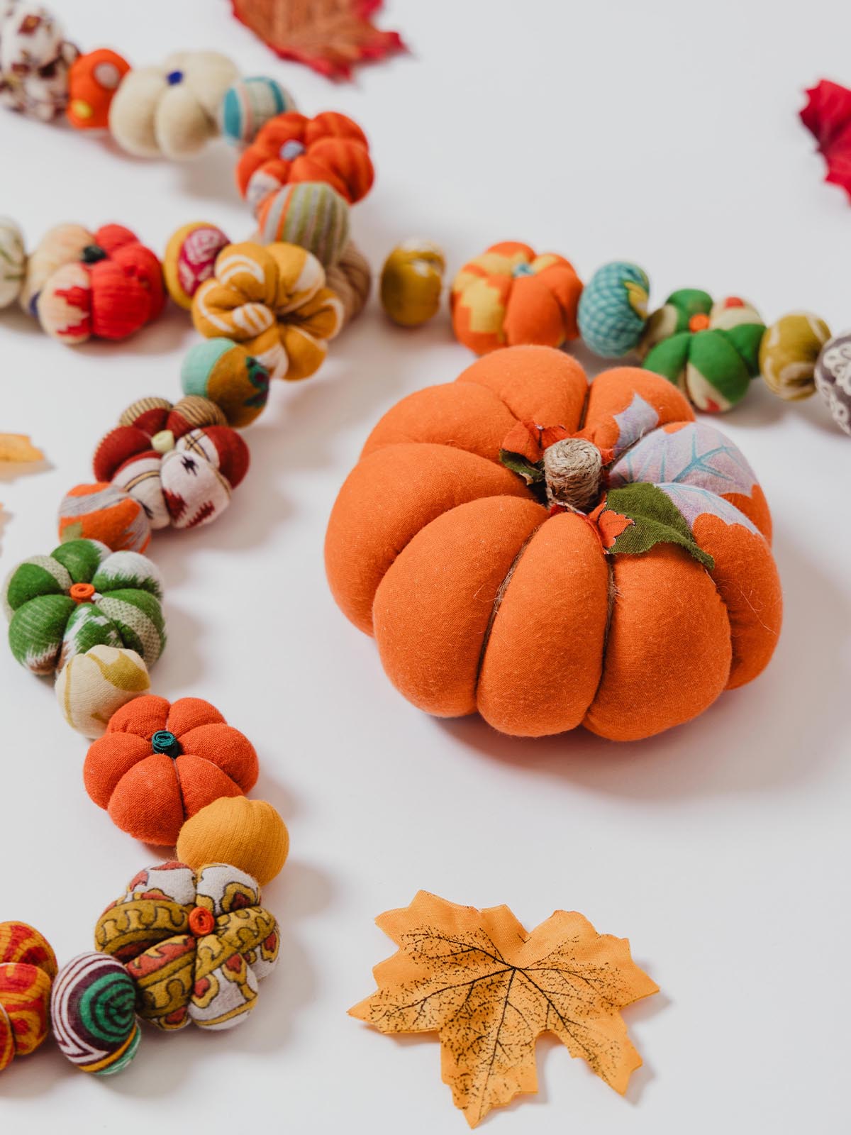 Close-up of an orange fabric pumpkin surrounded by a garland of miniature fabric pumpkins in assorted colors and patterns, with decorative fall leaves on a white background.