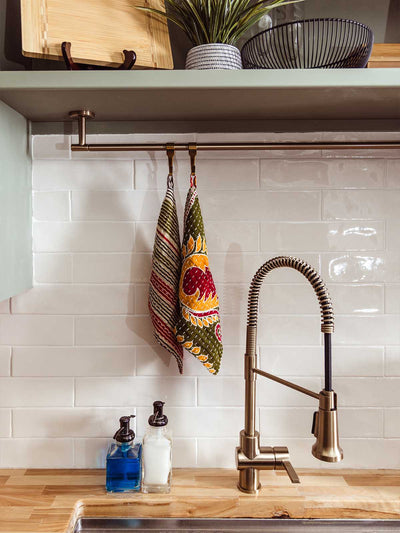 Handmade Kantha stitched kitchen towel with vibrant floral block print in green, orange, and red, hanging above farmhouse sink with modern brass faucet, eco-friendly artisan kitchen décor by Asha Project.
