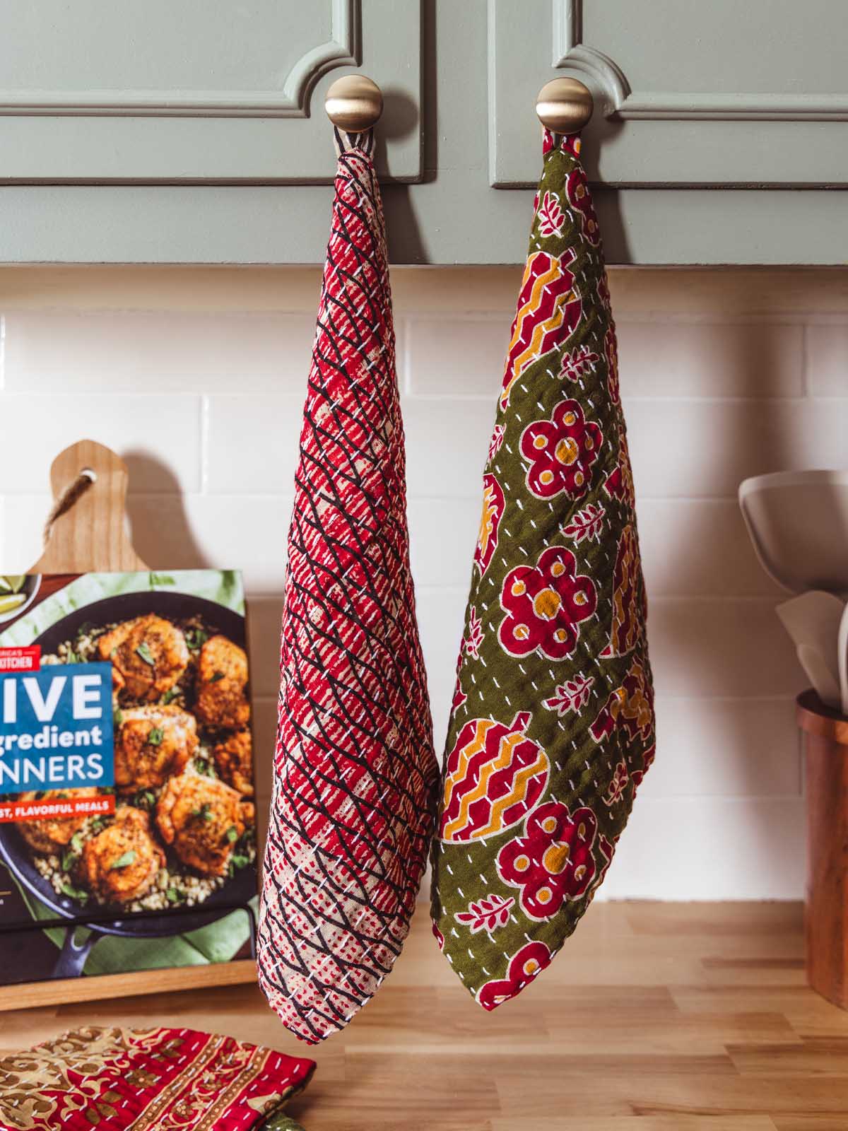 Two colorful kantha-stitched dish clothes hanging on kitchen cabinet knobs, featuring red and green block print patterns — handmade fair trade kitchen textiles.