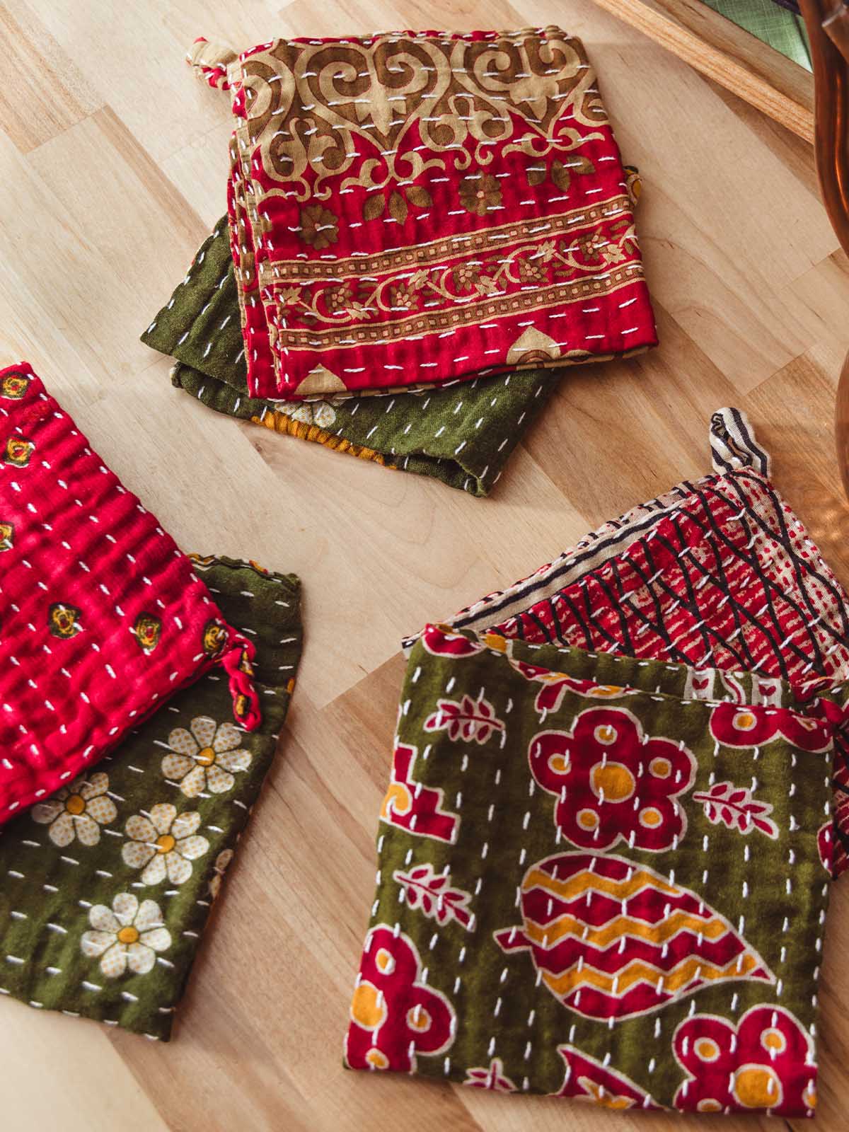 Stack of vibrant kantha-stitched dish clothes with red, green, and gold floral block prints on a wooden kitchen countertop — artisan-made sustainable kitchen décor.