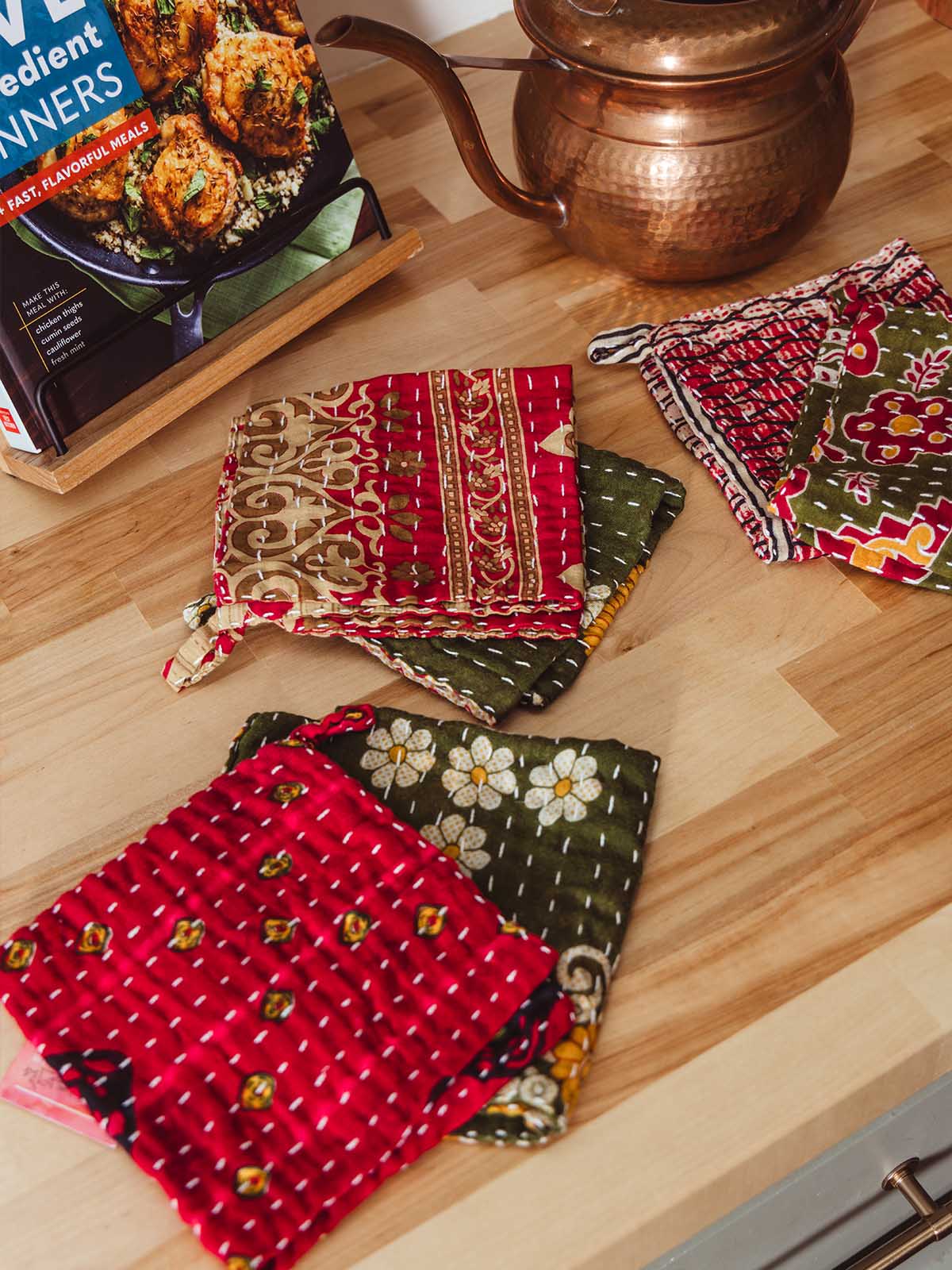 Assortment of handmade kantha dish clothes in rich red and green block print patterns styled on a wooden kitchen counter beside a copper kettle — fair trade kitchen accessories.