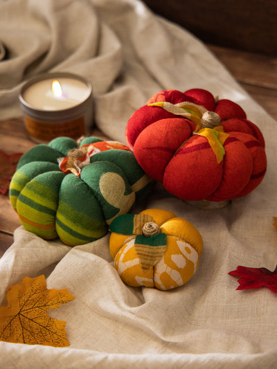 Three handmade fabric pumpkins in red, green, and yellow patterns displayed on a neutral linen cloth with a lit candle and decorative autumn leaves.