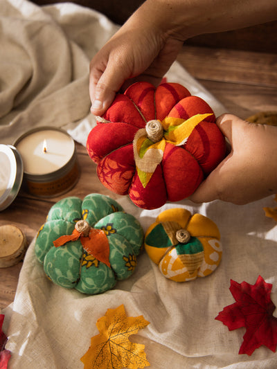 Hands holding a red handmade fabric pumpkin above a display of additional green and yellow pumpkins, a lit candle, and autumn leaves on a linen cloth.