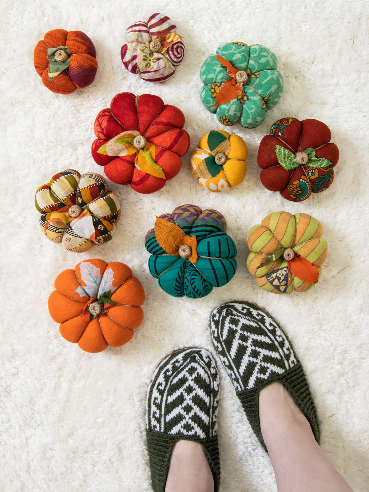 Overhead view of nine colorful handmade fabric pumpkins in various patterns and sizes arranged on a white textured surface beside feet wearing green and white knit slippers.