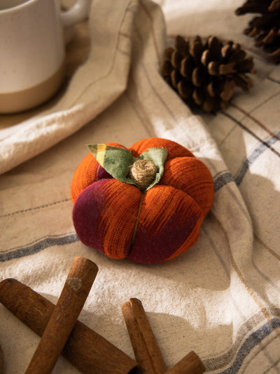Small orange and burgundy handmade fabric pumpkin on a striped neutral cloth with pinecones, cinnamon sticks, and a ceramic mug.
