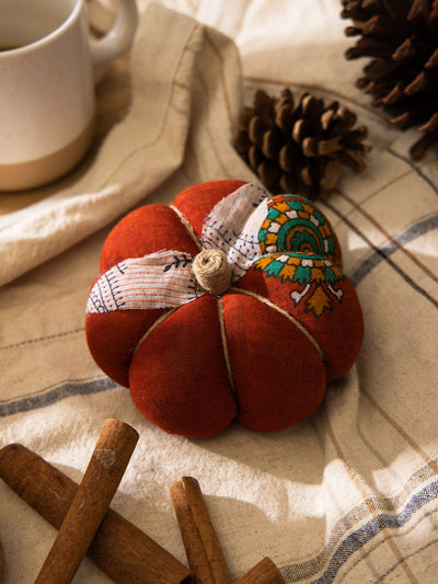 Burnt orange handmade fabric pumpkin with patterned fabric accents, placed on a striped cloth with pinecones, cinnamon sticks, and a ceramic mug.