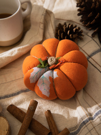 Large bright orange handmade fabric pumpkin with leaf-shaped fabric accents, displayed with pinecones, cinnamon sticks, and a ceramic mug.