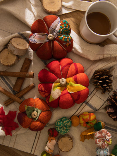 Overhead view of three handmade fabric pumpkins in orange, red, and burnt orange alongside a ceramic mug, cinnamon sticks, pinecones, wood slices, and a garland of miniature fabric pumpkins.