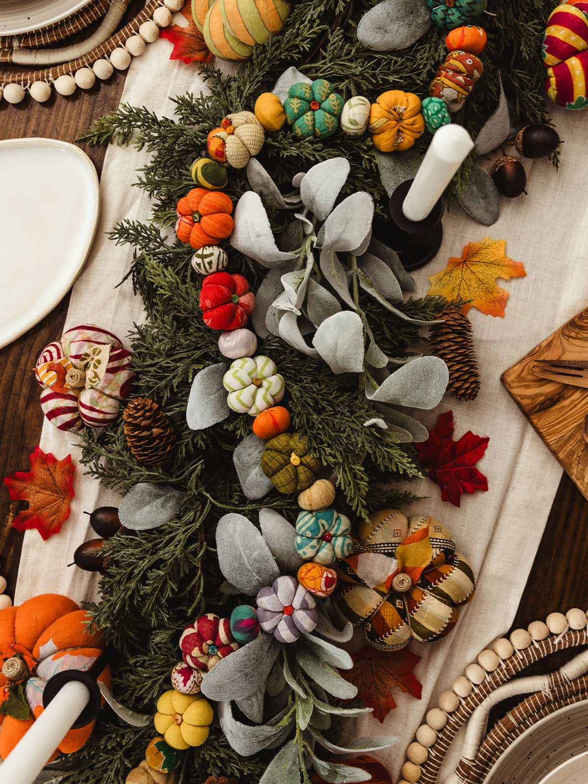 Festive fall table centerpiece featuring a long garland of colorful fabric pumpkins, greenery, pinecones, and faux lamb’s ear leaves, accented with autumn leaves and candles on a neutral table runner.