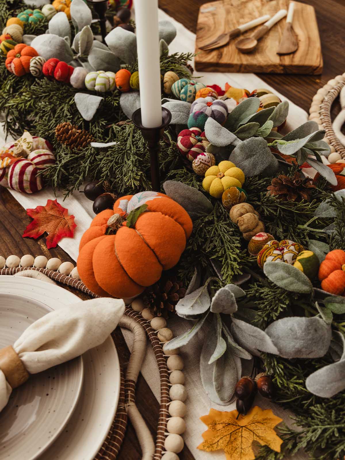 Side view of a fall tablescape with an orange fabric pumpkin, greenery garland, colorful fabric pumpkins, faux leaves, and taper candles, placed alongside a plate with a napkin and wooden bead charger.