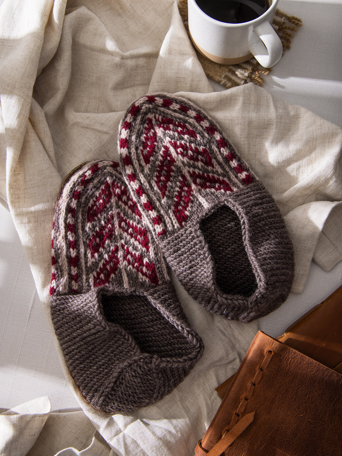 Pair of brown knit slippers with red and cream chevron and diamond patterns, styled on a beige linen cloth with coffee and a leather notebook.