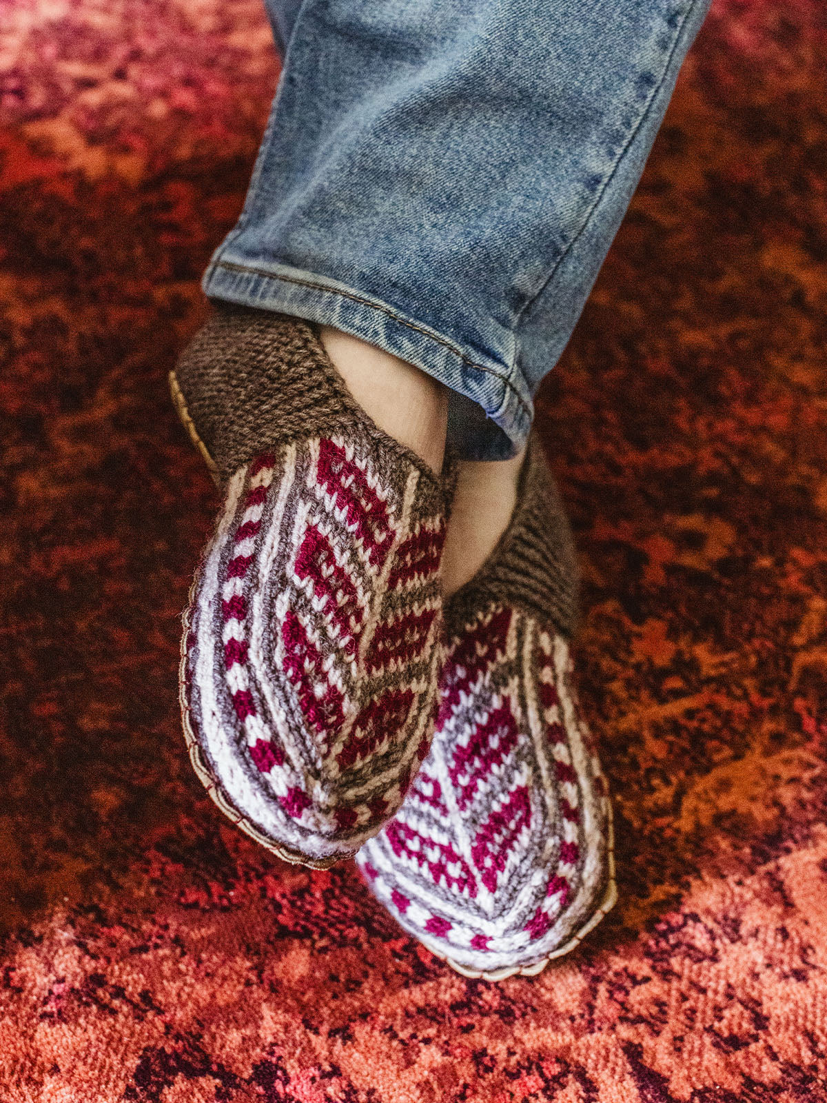 Male feet wearing red and tope colored handmade slipper socks on a red carpet.