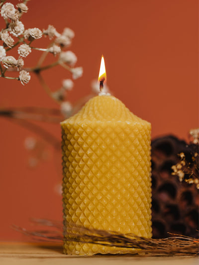 Lit beeswax candle with a textured pattern, casting a warm glow against a rich, orange background. Delicate white flowers and natural elements surround the candle, creating a cozy and inviting atmosphere.
