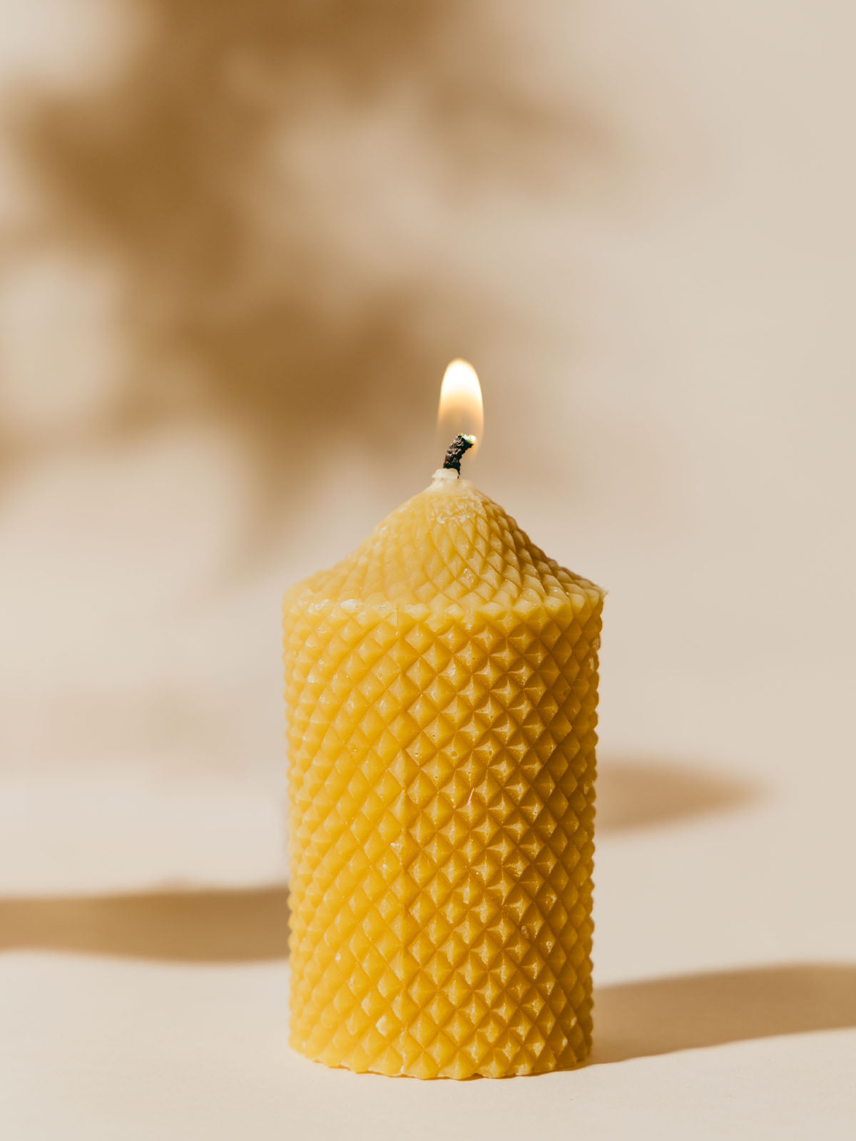 Beeswax candle with a diamond-patterned texture, lit and standing tall against a soft, neutral background. The simple and elegant design is accentuated by the warm light of the candle's flame