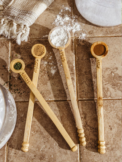 Flat lay of four carved wooden measuring spoons filled with spices and flour, styled on a tiled kitchen counter with a spilled dusting of flour and a fringed kitchen towel in the corner.