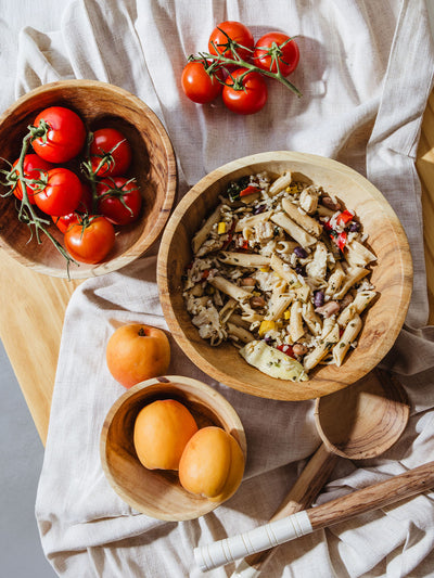 Overhead view of a rustic kitchen setup with a wooden bowl of penne pasta salad, a bowl of vine-ripened tomatoes, a bowl of apricots, and two wooden spoons arranged on a light linen cloth.