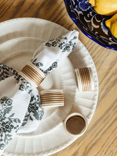 Set of woven cream and tan napkin rings on a white ceramic plate with a floral-patterned napkin threaded through one ring. A blue and white ceramic bowl filled with lemons sits nearby on a wood table.