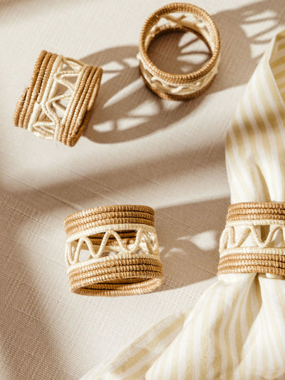 Set of four handwoven napkin rings crafted from natural fibers in tan and ivory, featuring a zigzag stitch pattern; one ring is shown in use around a striped fabric napkin, all styled on a light textile surface in warm sunlight.