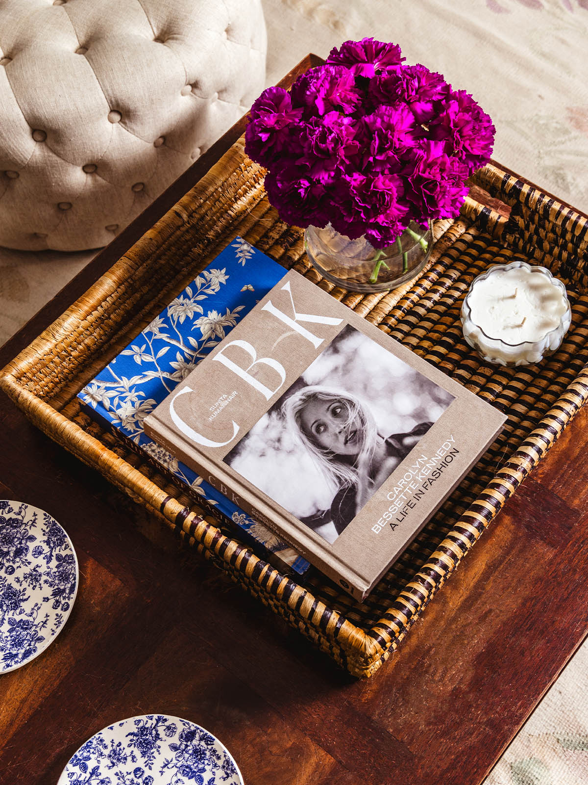 Cozy coffee table vignette featuring a woven tray with a Carolyn Bessette Kennedy fashion book, a vase of vibrant purple flowers, and a white decorative candle on a wood surface.
