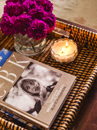 Close-up of a woven tray with a glowing candle, vibrant purple blooms in a glass vase, and the fashion book “Carolyn Bessette Kennedy: A Life in Fashion,” styled on a coffee table.