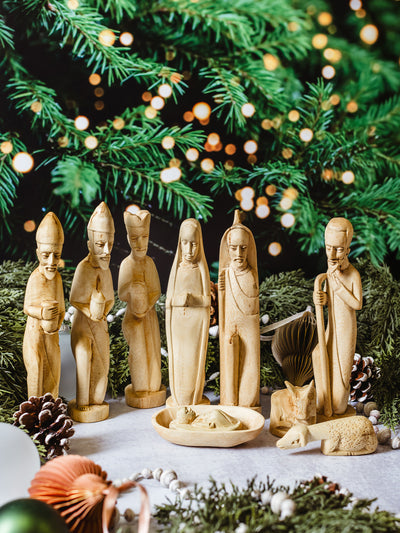 Full handcrafted wooden nativity scene with Mary, Joseph, baby Jesus, wise men, and animals styled with greenery, pinecones, and holiday lights — fair trade Christmas decoration.
