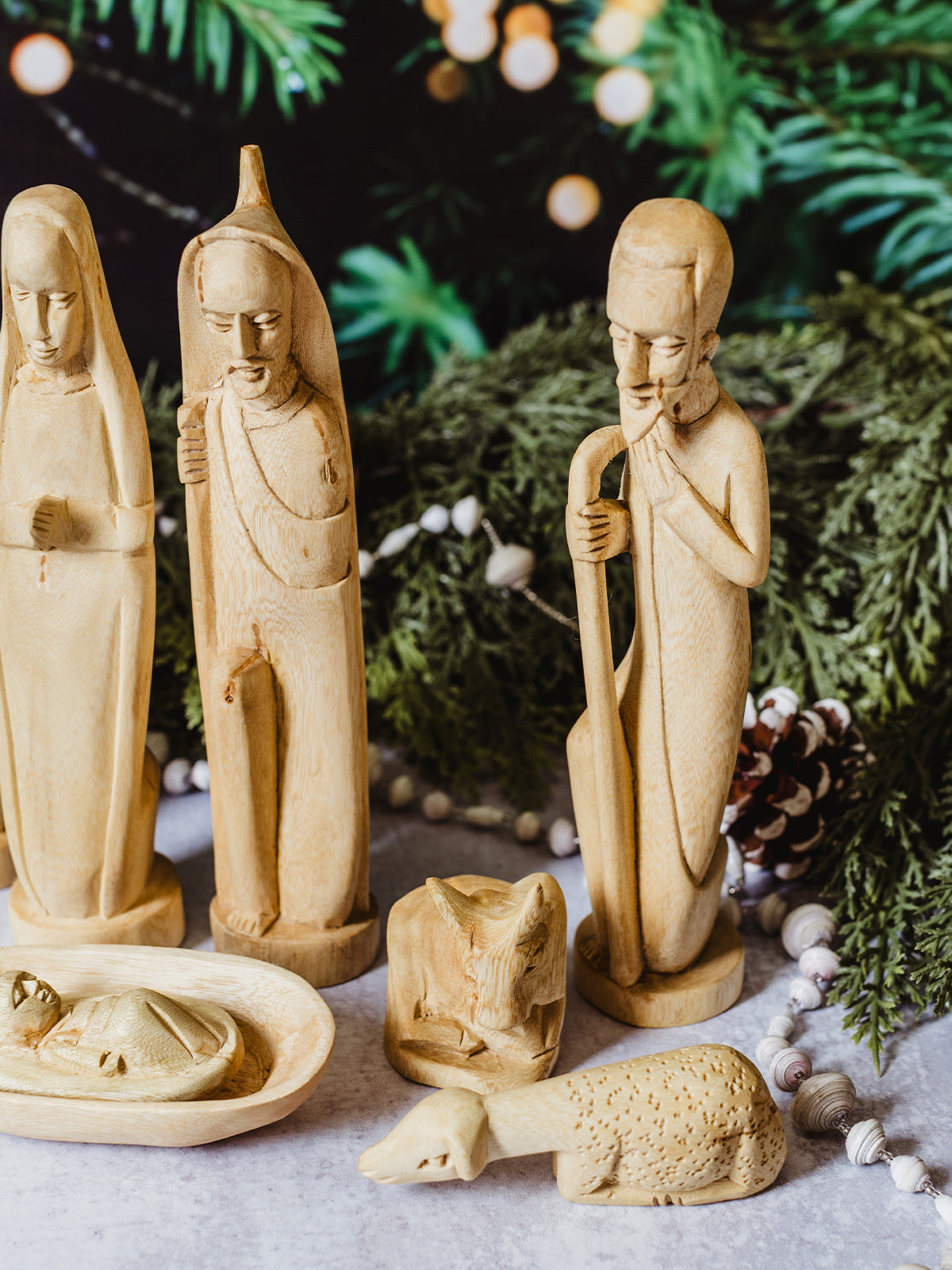 Close-up of carved wooden Joseph, Mary, and nativity animals with baby Jesus in a manger, surrounded by greenery and festive holiday accents — artisan-made Christmas décor.