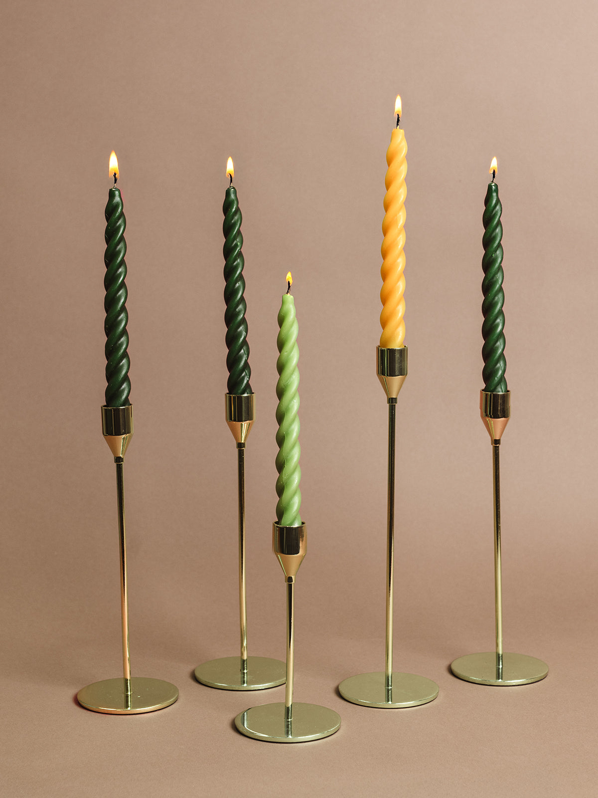 Five elegant twisted taper candles — forest green, soft sage, and gold — displayed in sleek brass candle holders against a neutral background. Ethically made home decor and sustainable holiday candle set.