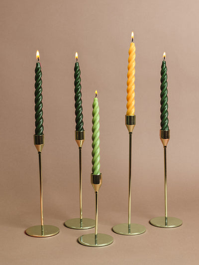 Five elegant twisted taper candles — forest green, soft sage, and gold — displayed in sleek brass candle holders against a neutral background. Ethically made home decor and sustainable holiday candle set.