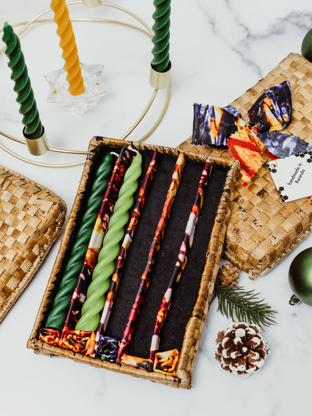 Gift box of fair trade handmade twisted taper candles in green and gold with patterned fabric wraps, displayed beside a brass Advent wreath and holiday greenery. Sustainable, ethical Christmas gift idea.