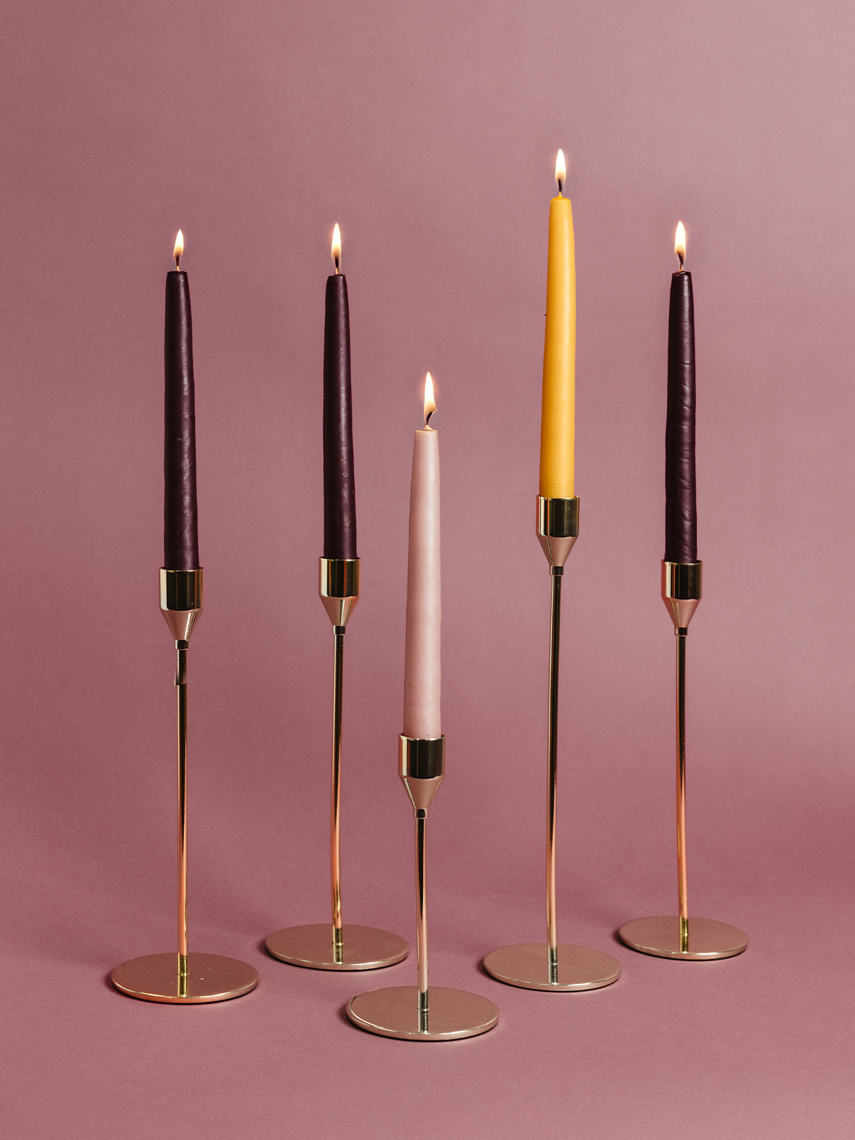 Five tall taper candles — rich purple, blush pink, and gold — glowing in modern brass candle holders on a rose background. Ethical Advent candle set for Christmas decor and gifting.