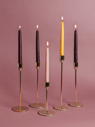 Five tall taper candles — rich purple, blush pink, and gold — glowing in modern brass candle holders on a rose background. Ethical Advent candle set for Christmas decor and gifting.