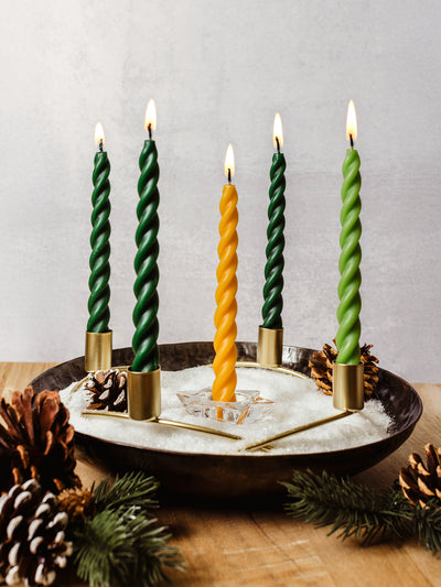 Five twisted taper candles — four deep green and one golden yellow — glowing in a modern Advent wreath with brass holders, faux snow, pinecones, and evergreens. Ethically made candles for holiday decor and sustainable Christmas traditions.