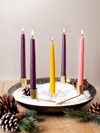 Set of five Advent taper candles — three deep purple, one pink, and one gold — burning in a minimalist brass wreath centerpiece with pinecones and faux snow. Handmade fair trade candles for Christmas Advent celebrations.
