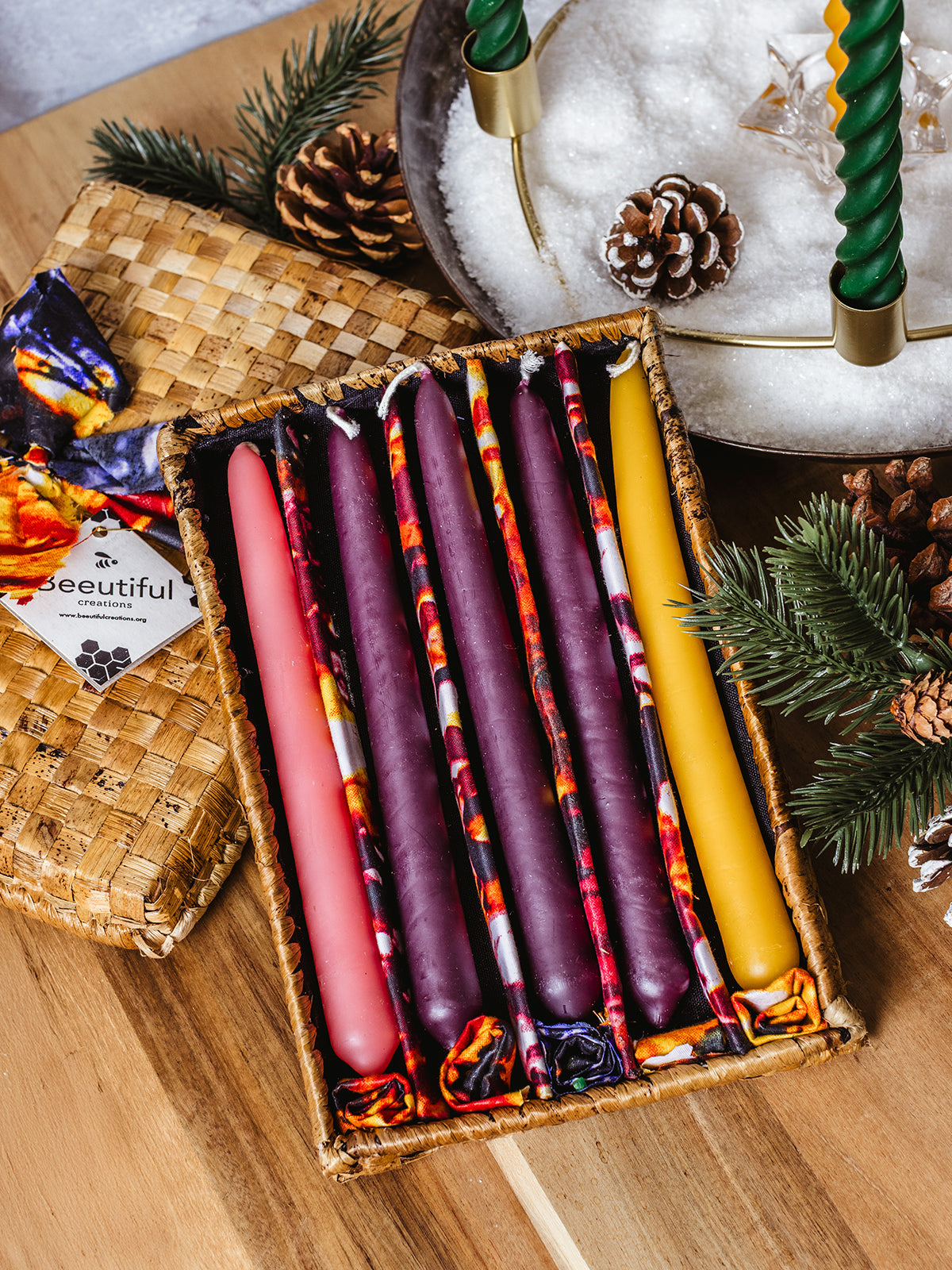 Handwoven basket filled with ethically made Advent taper candles — purple, pink, and gold — with colorful fabric wraps. Shown beside a brass holiday wreath centerpiece with pinecones and evergreen sprigs.