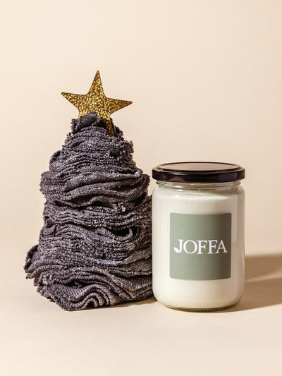 Handcrafted charcoal gray washcloth tree topped with a gold star, paired with a minimalist white candle in a glass jar labeled JOFFA, set against a warm neutral background.