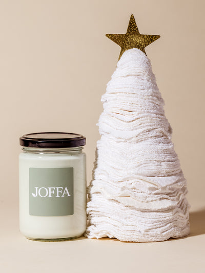 Handcrafted white washcloth tree topped with a gold star, shown next to a JOFFA-branded white candle in a clear glass jar, styled against a beige backdrop.