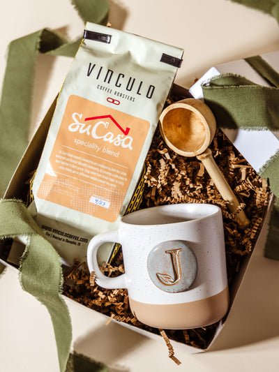 Flat lay of a Vinculo Su Casa speciality blend coffee bag, “J” monogram mug, and wooden scoop arranged in a gift box with brown crinkle paper and green ribbon, perfect for thoughtful corporate gifting.