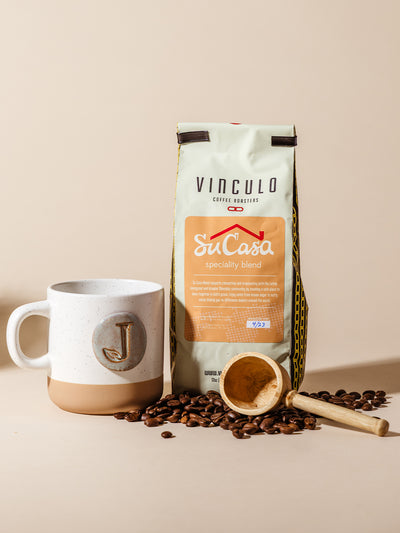 Vinculo Su Casa speciality coffee bag displayed with scattered beans, a handcrafted “J” ceramic mug, and wooden scoop, styled on a neutral background to highlight artisan detail and cozy gifting appeal.