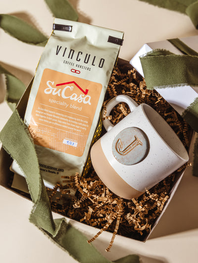 Flat lay of Vinculo Su Casa speciality blend coffee and personalized “J” ceramic mug inside a gift box with brown crinkle paper and green ribbon, styled for thoughtful corporate gifting.