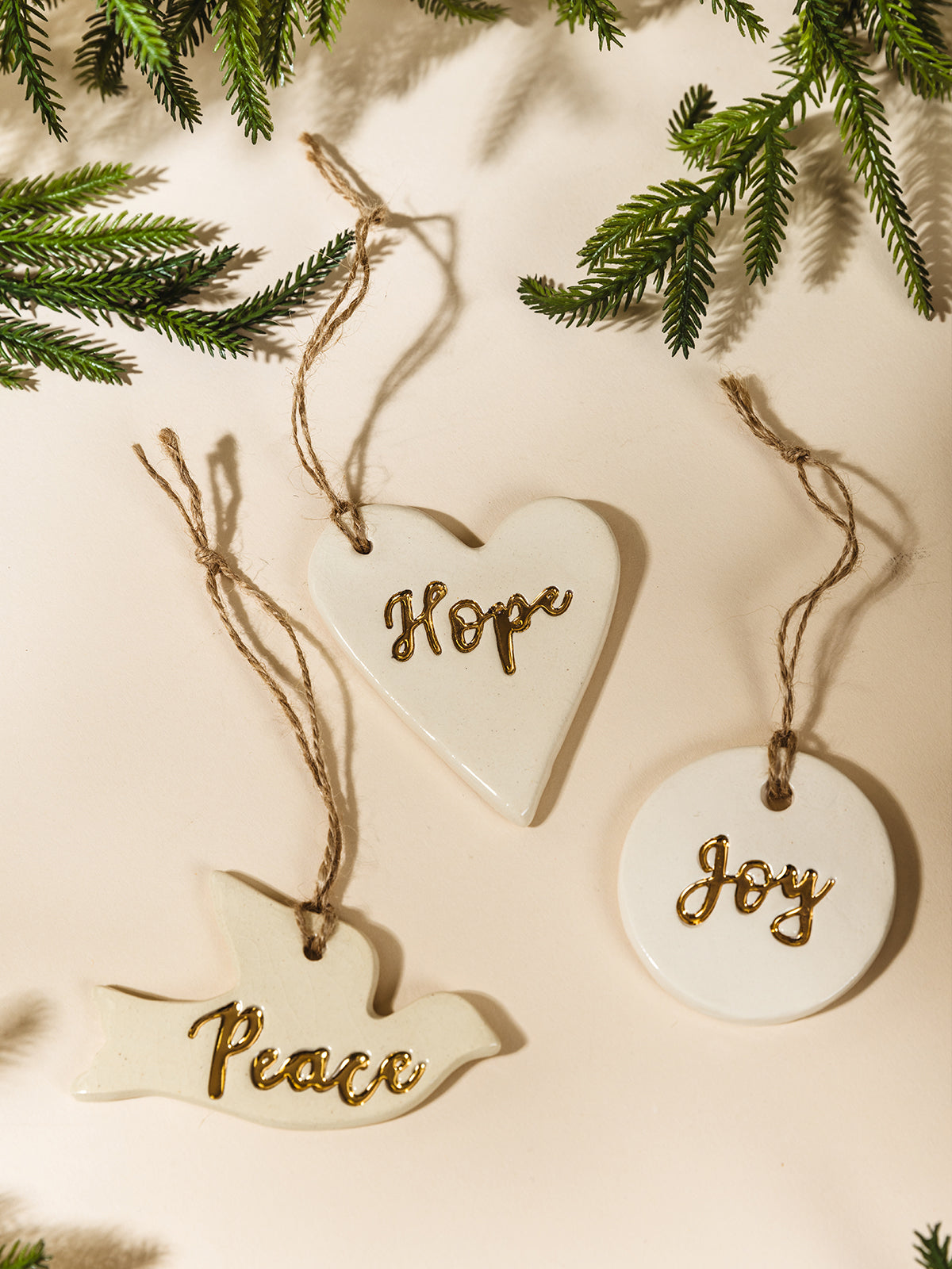 Three handcrafted ceramic ornaments—heart with “Hope,” circle with “Joy,” and dove with “Peace”—each with gold lettering and twine hangers, styled flat with evergreen branches for a festive holiday feel.