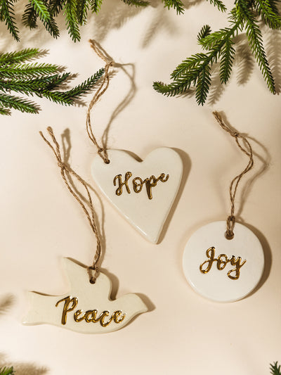 Three handcrafted ceramic ornaments—heart with “Hope,” circle with “Joy,” and dove with “Peace”—each with gold lettering and twine hangers, styled flat with evergreen branches for a festive holiday feel.