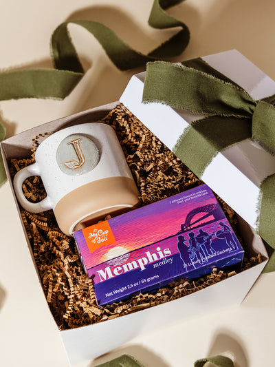 Flat lay of a white gift box filled with crinkle paper, featuring a Memphis Medley tea sampler by My Cup of Tea and a handcrafted “J” ceramic mug, tied with an olive green ribbon for a polished, locally inspired corporate gift.