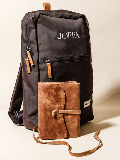 Sleek black backpack with JOFFA logo and tan leather zipper pulls, paired with a rustic brown leather wrap journal, styled upright against a neutral background for a modern and practical gift pairing.