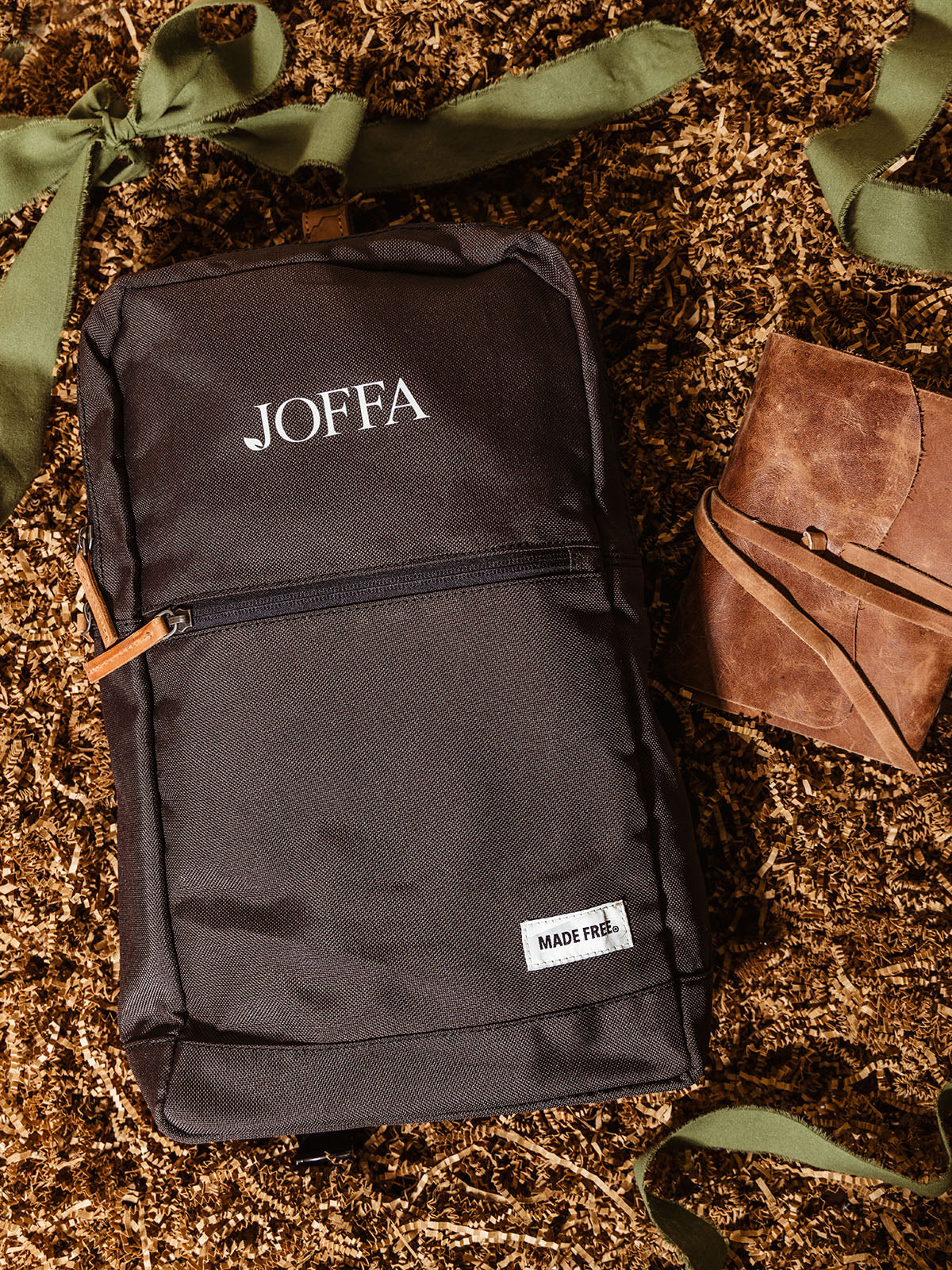 Flat lay of JOFFA-branded backpack and leather wrap journal placed on kraft crinkle paper with green ribbon, styled as an elevated and meaningful corporate gift with impact-driven design.